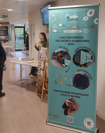 Stand d'animation et sensibilisation au compostage et tri des déches alimentaires en ville animé par OuiCompost Un stand de OuiCompost animé par une animatrice de OuiCompost. Un grand kakémono avec le logo de OuiCompost et les images de collecte des biodéchets est posé devant le stand. Le public près du stand écoute les explication de l'animatrice.