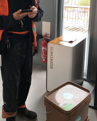 Une poubelle métallique avec l'inscription "biodéchets" sur la porte. Un collecteur de OuiCompost à côté de la poubelle est en train de réaliser la collecte de biodéchets à Lyon et rentrer les données de la collecte dans la base de données OuiCompost.