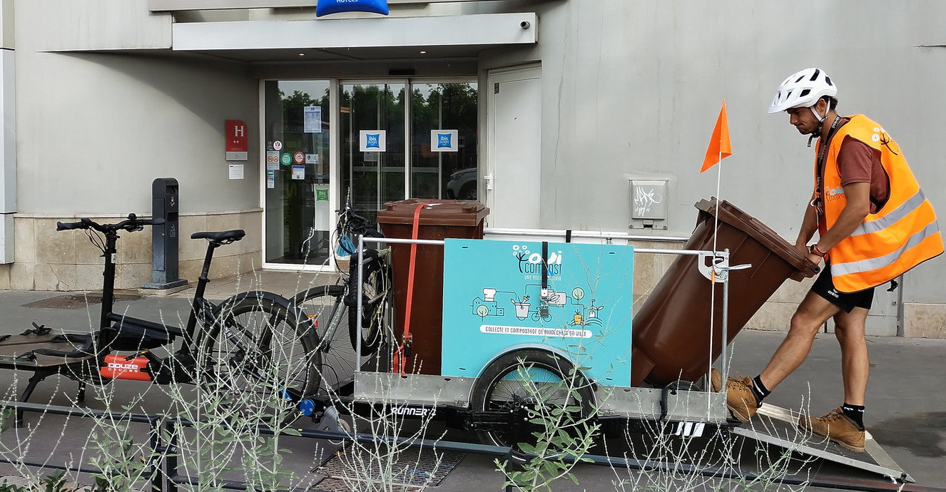 Collecte de bacs de 120 litres par OuiCompost Un collecteur de OuiCompost décharge les bacs roulants de 120 lires d'une remorque de vélo pour réaliser une collecte de biodéchets par échange de bacs. La remorque est floquée Ouicompost et possède une rampe pour descendre et charger les bacs roulants.