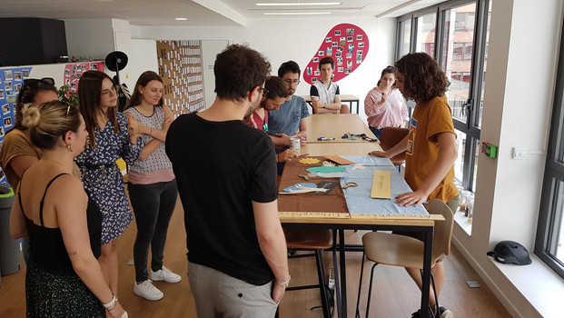 Une séance de sensibilisation de compostage animée par OuiCompost dans les bureaux d'un client. Un groupe de collaborateurs se tiennent autour d'une table pour écouter les explications à propos du tri des déchets alimentaires et le compostage urbain. Sur la table, on voit les photos et les jeux ludiques proposés aux participants de l'atelier. 