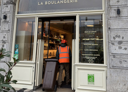 Collecte de déchets organiques à vélo dans les commerces de Lyon Agent de collecte en gilet orange transportant une poubelle roulante à biodéchets dans une boulangerie à Lyon dans le cadre d’un service de collecte de déchets organiques à Lyonà vélo.