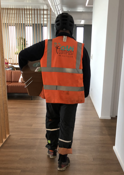 Un collecteur de OuiCompost rentre dans les bureaux d'un client pour collecte les déchets alimentaires de bureau. Le collecteur de OuiCompos porte un gilet orange avec un grand logo de OuiCompost sur le dos et transporte un seau à biodéchets pour remplacer un bac plein qui s'apprête à collecter.. 