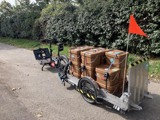 Un vélo cargo avec une remorque est chargé des petits bacs à biodéchets que OuiCompost utilise pour la collecte de déchets organiques à Lyon. Les bacs marrons de 22 litres sont fixés sur la remorque avec les sangles. Le chargement est prêt pour la collecte.