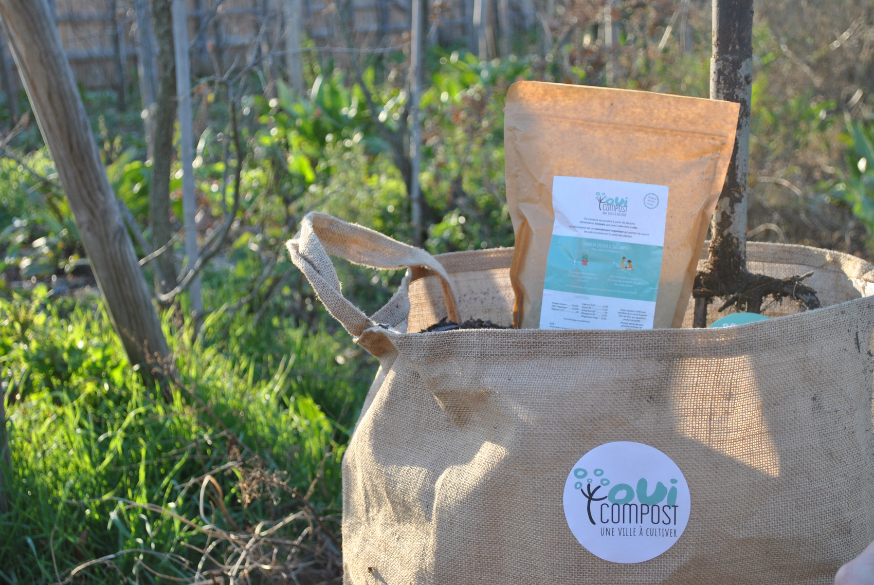 Un petit pochon de compost est posé dans un grand sac en jute. Une fourche est plantée dans le grand sac et on voit de la verdure sur à l'arrière plan. Sur le pochon est collée une étiquette avec le logo de OuiCompost et les information sur le compost issu des collectes des déchets organiques à Lyon. Un grand logo OuiCompost est collé sur le sac en jute avec la phrase "Une ville à cultiver"  